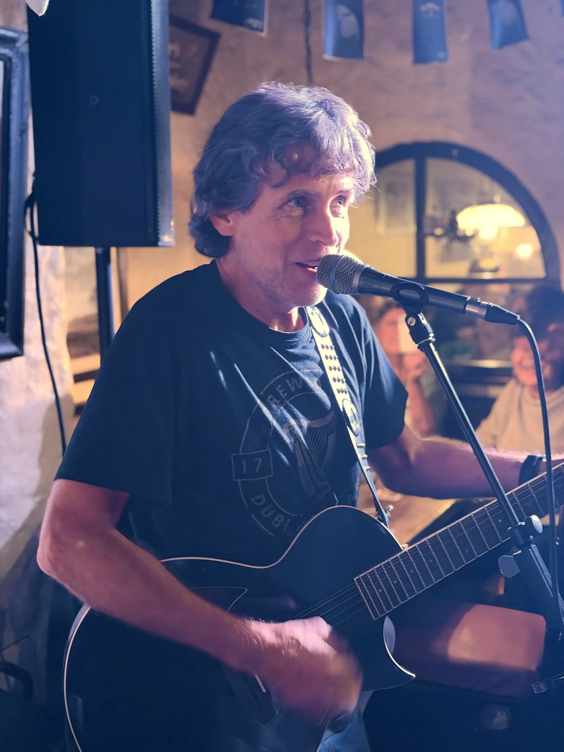 Walt lächelt am Mikrofon, mit schwarzer Akustikgitarre, im Hintergrund ein Rundbogenfenster im Pub